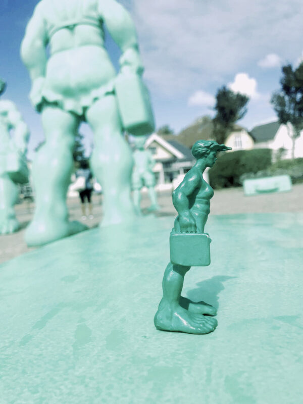 Die 10 cm große Miniatur der grünen Sylter Riesin steht auf dem grünen Koffer vor den original Skulpturen der "Reisenden Reisen im Wind" auf dem Westerländer Bahnhofsvorplatz. Von Neptunsgeschmeide und Martin Wolke.