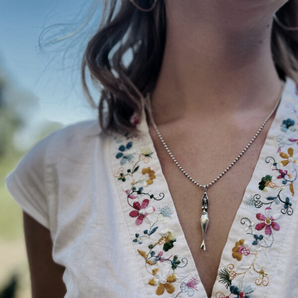 Ein Meerjungfrau-Anhänger von Neptunsgeschmeide hängt an einer Silberkette am Dekolletee einer jungen Frau mit Bluse und Blumenkragen.