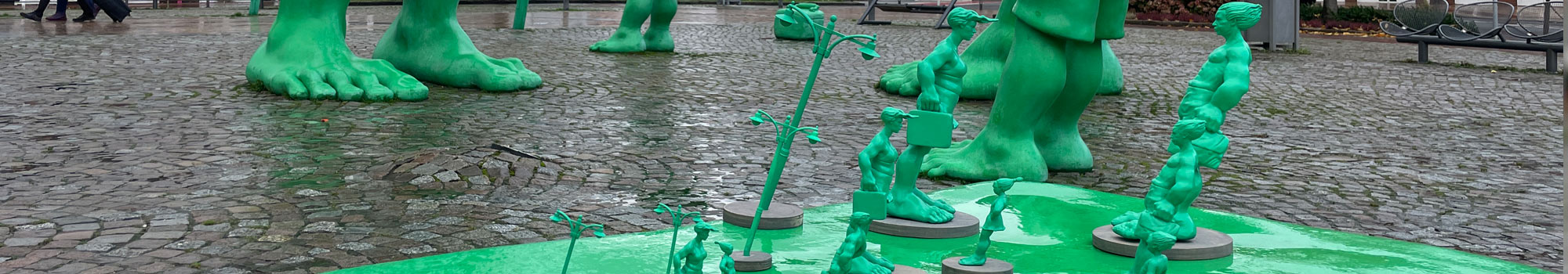 Reisende Riesen im Wind, Skulpturen von Martin Wolke. Alle Miniaturen in Größen von 10 - 30 cm stehen auf dem echten grünen Koffer vor den Riesen auf dem Westerländer Bahnhofsvorplatz auf Sylt. 