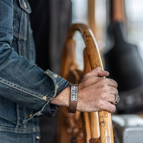 Koordinaten Armband von Neptunsgeschmeide. Ein Hand-geprägtes Zinnplättchen ist auf einem Lederarmband mit Knopfnietenverschluss vernietet. Abbildung zeigt die Koordinaten von List auf Sylt. Bild zeigt eine Hand, die auf der Brücke das Steuerrad hält.