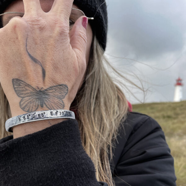 Koordinaten Armreif von Neptunsgeschmeide. Getragen von einer Frau vor dem Gesicht, mit den Koordinaten von List auf Sylt. Hintergrund zeigt den Sylter Leuchtturm auf dem Ellenbogen auf Sylt.