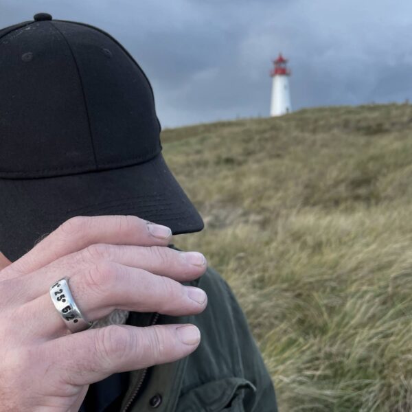 Vor einem Leuchtturm trägt eine Männerhand einen Koordinaten Ringe von Neptunsgeschmeide. Hand geprägt und in Silberzinn gegossen.