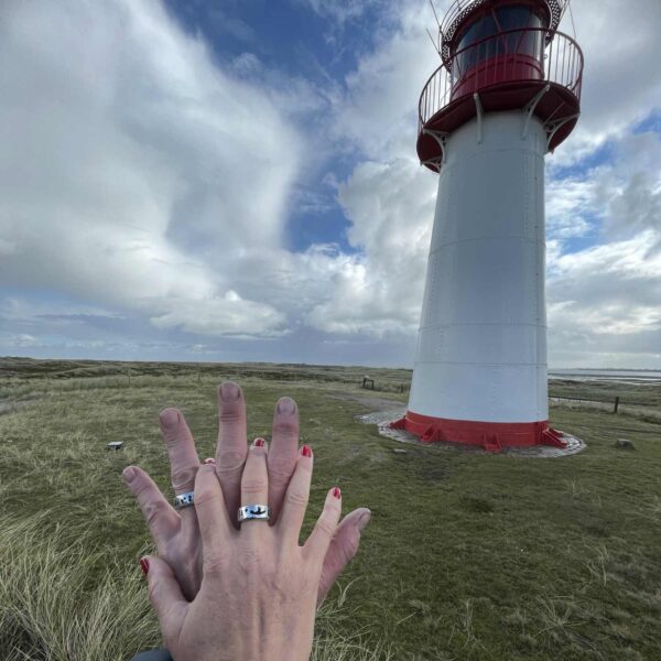 Ein Paar hält sich vor einem Sylter Leuchtturm die Hände und trägt die Sylt Koordinaten Ringe von Neptunsgeschmeide. Hand geprägt und in Silberzinn gegossen.