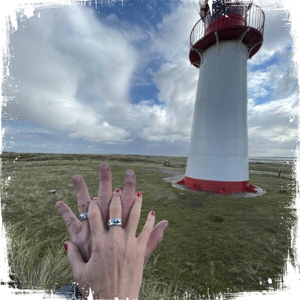 Ein Paar hält sich vor einem Sylter Leuchtturm die Hände und trägt die Sylt Koordinaten Ringe von Neptunsgeschmeide. Hand geprägt und in Silberzinn gegossen.